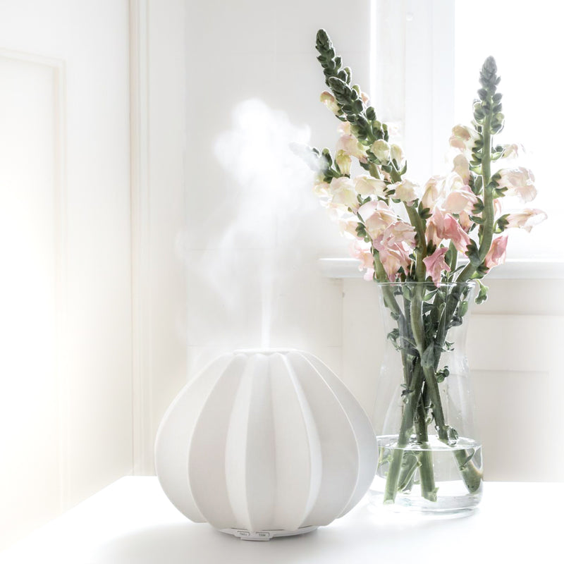 Der Aroma Diffuser aus weisser Keramik mit Dampf vor einer weissen Wand mit Struktur und daneben eine Gläserne Blumenvase mit Blumen (Löwenmäulchen) drin. 